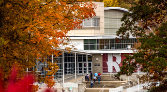 Image shows students walking up the front steps of the Sports and Recreation Center where a big Rose-Hulman R is on the exterior wall. The image is framed on the right and left by colorful leaves of two trees.