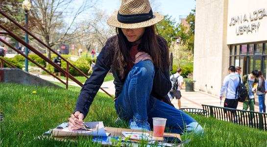 Female student sit in the grass in Root Quadrangle doing watercolor drawing.