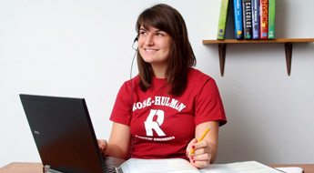 Image shows a female student smiling and looking off to her right while wearing a red Rose-Hulman T-shirt and taking notes as she uses her laptop computer.