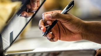 Image shows a student’s hand holding a pen next to a laptop computer. The student is taking notes while working.