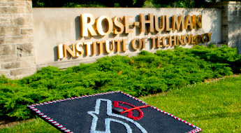 Image shows the large stone and concrete entrance sign for Rose-Hulman. The sign has large gold-colored letters reading Rose-Hulman Institute of Technology. The sign is lighted by bright sunlight and sits on green grass with low green bushes directly in front of the sign