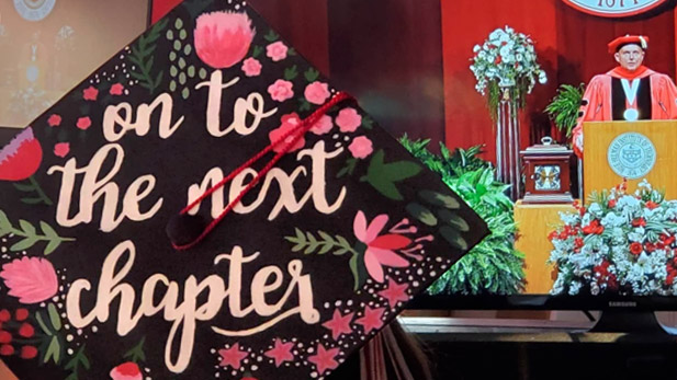 Top of a decorated student graduation cap reading: "Onto the Next Chapter." President Coons in regalia is at the podium on a screen in the distance.