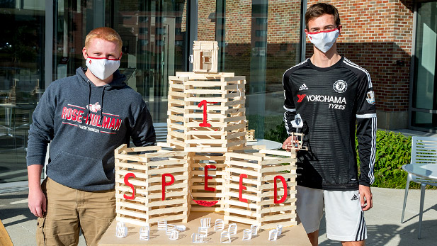 Two students standing with a bonfire model. The model includes lettering saying "Speed 1."