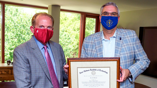 President Rob Coons presenting a framed Honorary Degree to Indiana Gov. Holcomb. Both men are wearing masks due to COVID-19 pandemic.