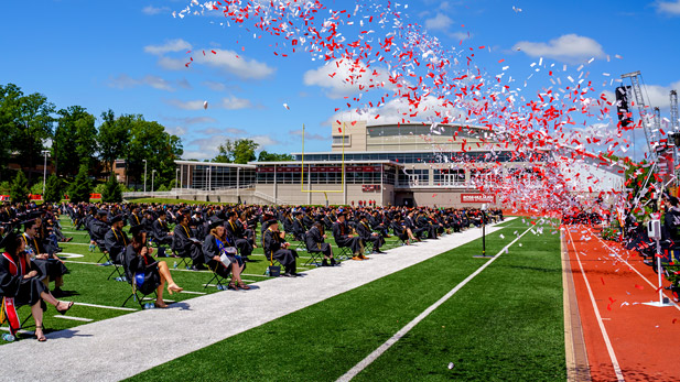 Graduates sit on the football field as red and white confetti rains down.
