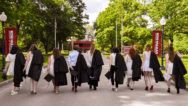 From-behind-view of several graduating seniors walking up the main road onto campus with their commencement regalia draped over their shoulders with Hadley Hall in the distance.