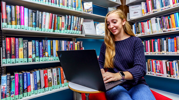 A student works on a computer near the bookshelves in the Logan Library.