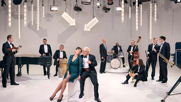Pink Martini Orchestra, wearing dresses and tuxedos, pose with their instruments.