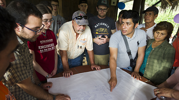 A group of people discuss a blueprint on a table, which one person is pointing to.