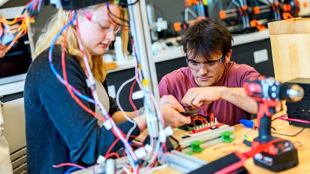 Two students wearing safety goggles and surrounded by equipment test the wires of their project.