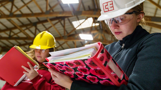 Two civil engineering students in hard hat write notes on their binders.