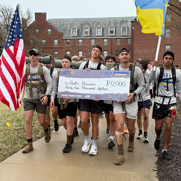 Group of Alpha Tau Omega members standing together holding a large check and their fraternity flag in front of Riley Hospital for Children.