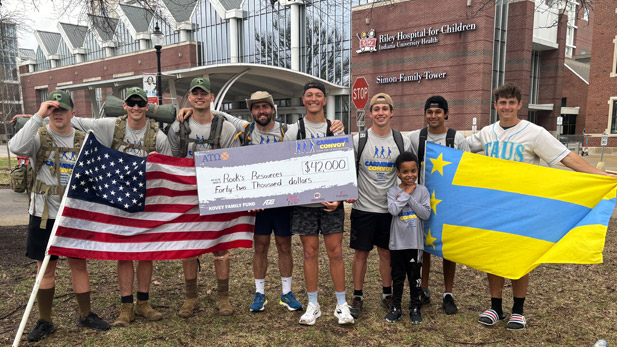 Group of Alpha Tau Omega members standing together holding a large check and their fraternity flag in front of Riley Hospital for Children.