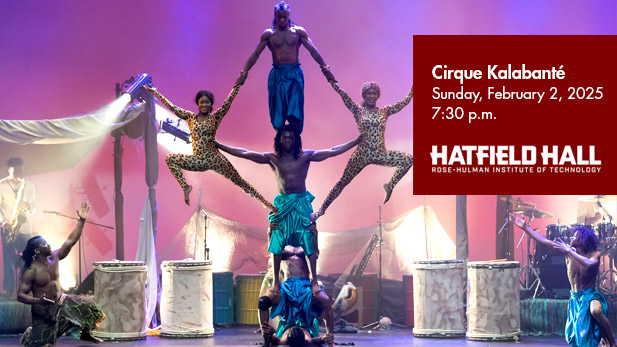 Six Cirque Kalabanté performers, wearing brightly colored costumes, balance on each others' bodies to perform an acrobatic stunt onstage. 