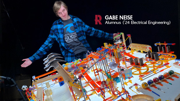 Gabe Neise stands proudly behind one of his machines, which contains several hammers and billiards balls, K'NEX pieces, dominoes, and other small household objects.