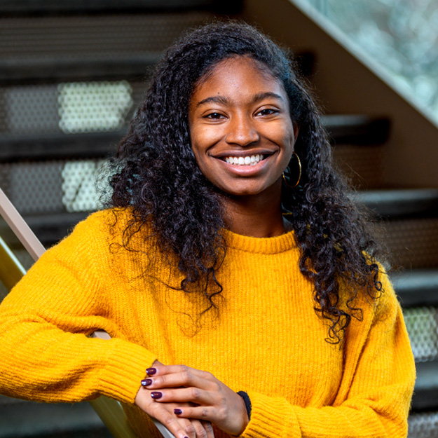 Janaé Gillus, a young Black female student wearing a yellow sweater, smiles in front of a wood paneled wall.