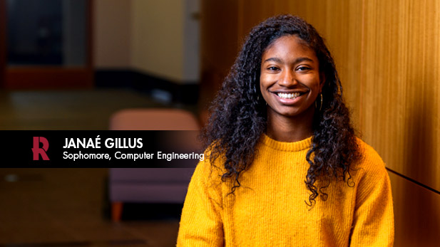 Janaé Gillus, a young Black female student wearing a yellow sweater, smiles in front of a wood paneled wall.