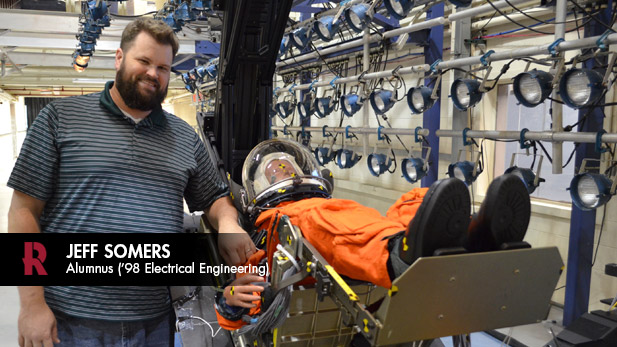 Jeff Somers poses beside a test mannequin in a spacesuit lying on an equipment prototype.