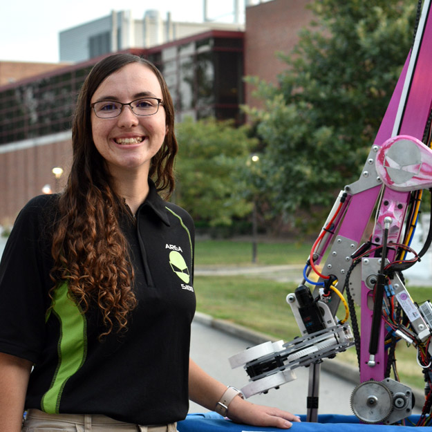 Katie Collins, wearing a black and green polo with Area 5188's alien logo, smiles next to the team's hot pink robot in front of the Branam Innovation Center.