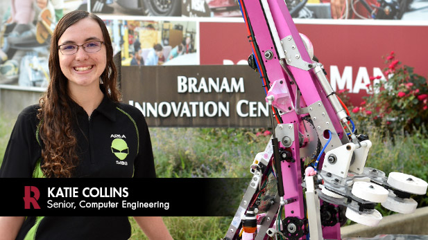 Katie Collins, wearing a black and green polo with Area 5188's alien logo, smiles next to the team's hot pink robot in front of the Branam Innovation Center.