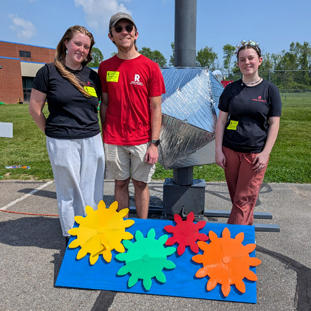 Two smiling volunteers pose enthusiastically beside a reflective geometric structure mounted on a pole, surrounded by colorful gear-shaped cutouts on the ground.
