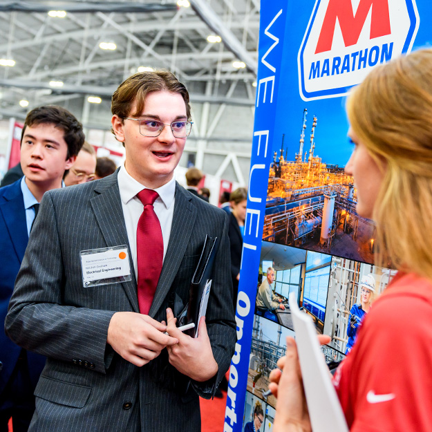 A student in professional clothing holds a padfolio while talking to an employer.