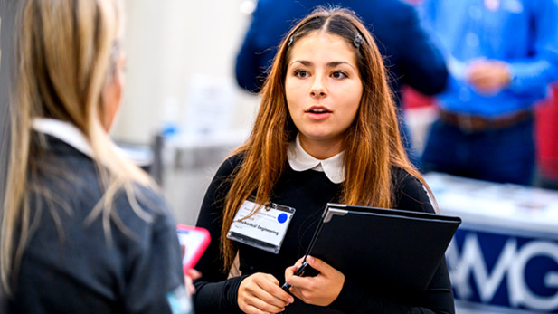 A student in professional clothing holds a padfolio while talking to an employer.