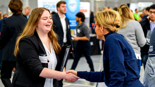 A student in professional attire smiles and shakes the hand of an employer.