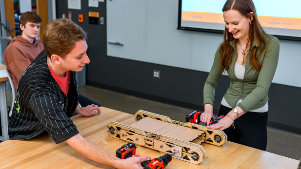 A faculty and students work together in a classroom lab, assembling and testing a cardboard prototype device on a table.