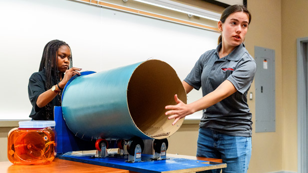 Two students present their project, which includes a large cylinder on wheels, at Engineerathon.