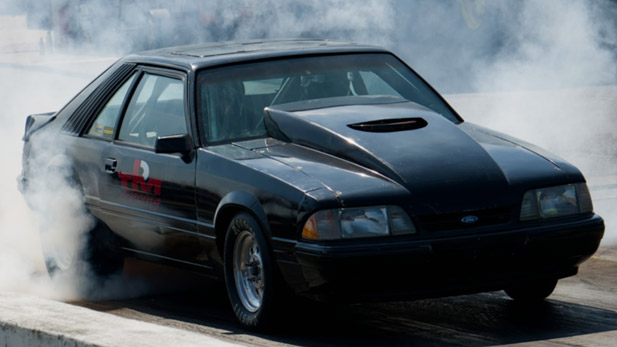 A black car with the Team Rose Motorsports logo speeds through a cloud of smoke.